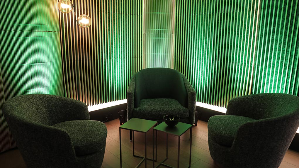Coin fauteuils avec trois fauteuils, tables basses et éclairage vert sur un mur à lattes verticales.