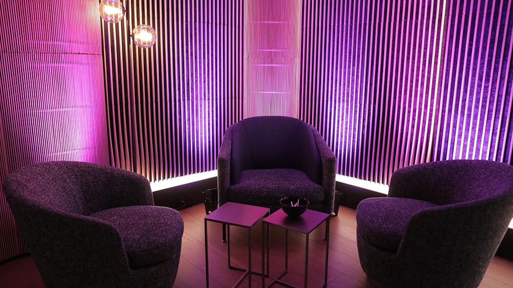 Coin fauteuils avec trois fauteuils, tables basses et éclairage rose et violet sur un mur à lattes verticales.