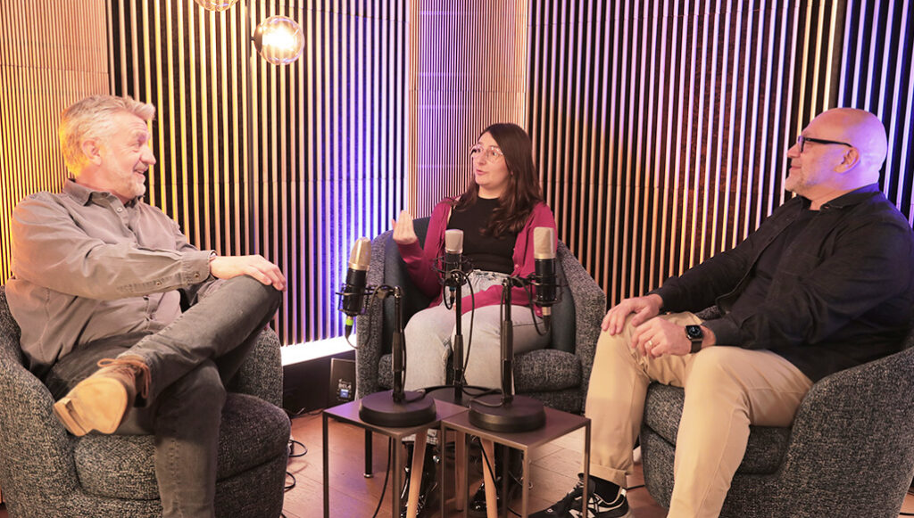 Trois personnes assises dans des fauteuils autour de microphones, discutant dans un studio de podcast chaleureux éclairé en tons dorés et violets.