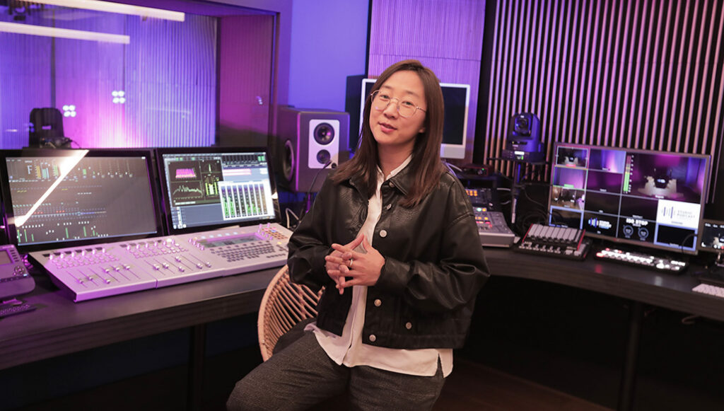 Personne assise dans une régie audio-vidéo moderne, entourée d’écrans de mixage, de contrôleurs et de moniteurs, éclairée par des lumières violettes et bleues.