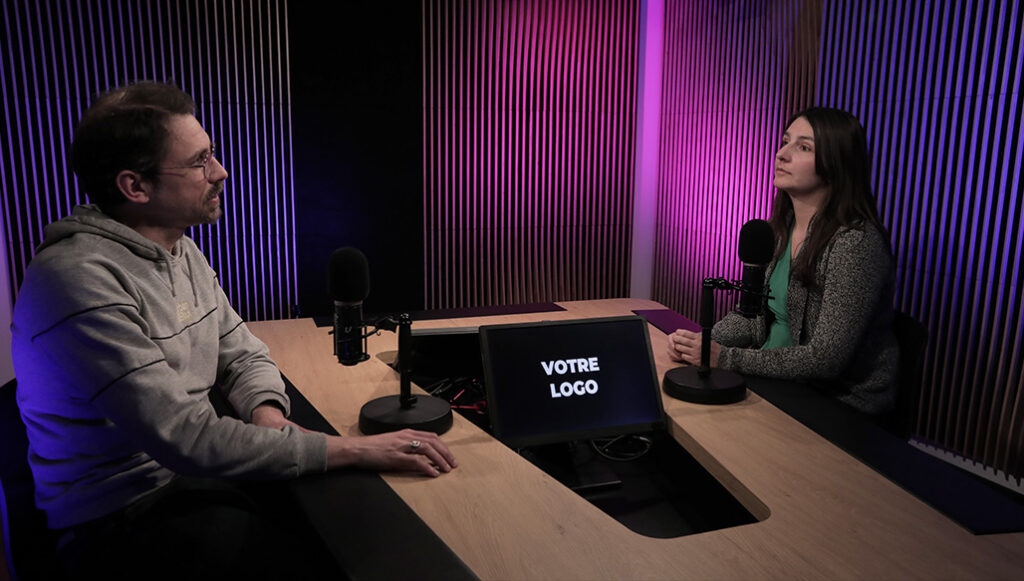 Deux personnes assises face à face dans un studio de podcast moderne, équipées de microphones professionnels et éclairées par des lumières violettes et bleues, avec un écran central affichant “Votre logo”.