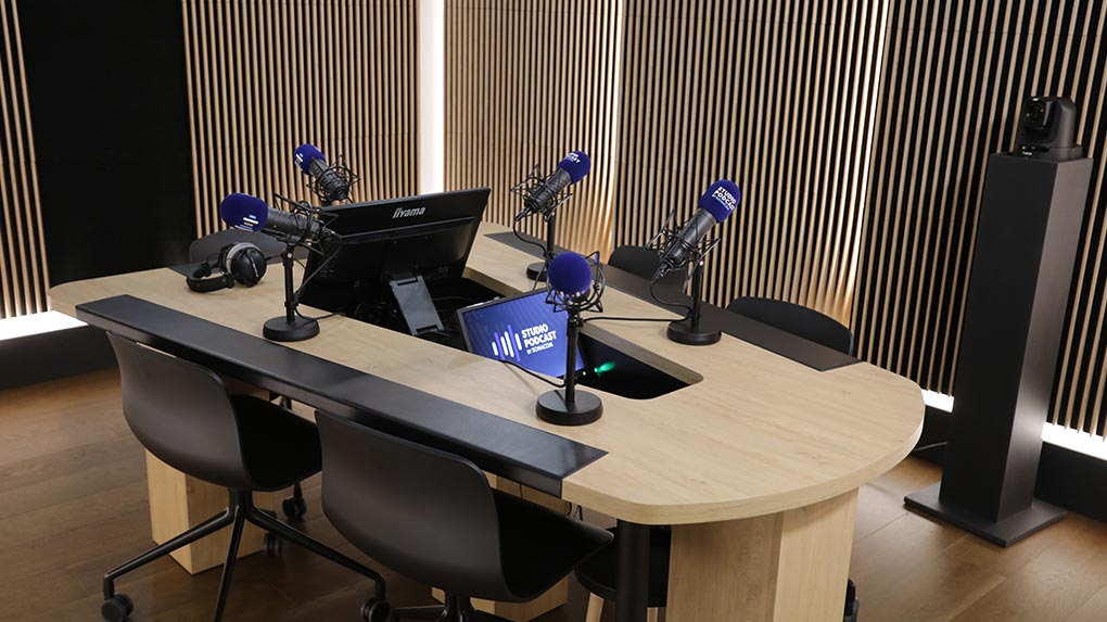 Studio de podcast professionnel avec table en bois, cinq micros, éclairage neutre et écran central personnalisable.